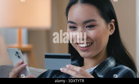 Femme asiatique joyeuse ethnique tenant une carte bancaire et un téléphone portable faisant le paiement en ligne achat Internet souriant coréen Japonais Chinois e-commerce féminin Banque D'Images