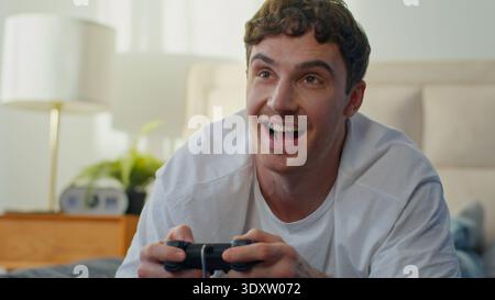 Heureux caucasien jeune homme couché au lit à la maison jouant au joystick de jeu vidéo souriant gars appréciant le jeu de console amusant passe-temps masculin mains tenant le contrôleur Banque D'Images
