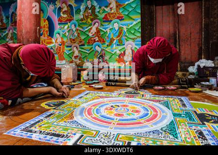 Les moines en robes marron et masques faciaux travaillent sur un mandala de sable détaillé avant de réaliser des peintures murales bouddhistes vibrantes à l'intérieur du Hemis Gompa. Banque D'Images