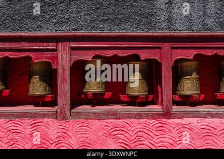 Gros plan de roues à prières dorées avec des inscriptions tibétaines placées dans un cadre en bois rouge contre un mur gris texturé à Thiksey Gompa. Banque D'Images