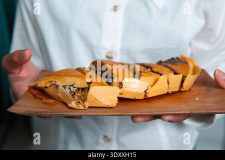 Vue latérale rapprochée de Martabak Manis tranché sur une assiette en bois, montrant une texture épaisse et une garniture de chocolat fondu sur un fond à motifs. Banque D'Images