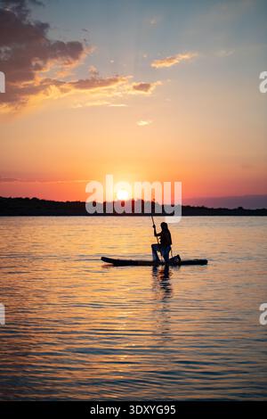 Silhouette d'une personne debout sur une planche à paddleboard au coucher du soleil, avec le coucher du soleil derrière une île. Ciel spectaculaire et eau calme. Banque D'Images