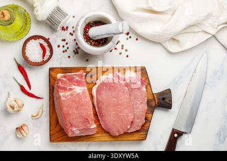 Porc. Viande crue fraîche sur une planche à découper, sel, poivre dans un mortier, ail, huile d'olive et piment rouge sur fond de marbre. Côtelettes de porc. Selecti Banque D'Images