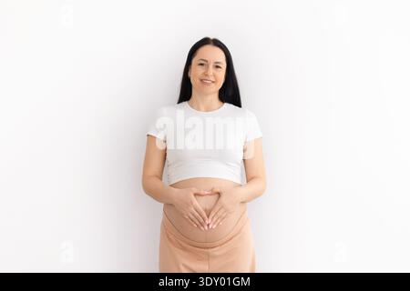 Jeune femme enceinte formant une forme de coeur avec les mains sur le ventre à la maison Banque D'Images