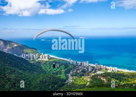 Parapente survolant l'océan Atlantique et la plage de São Conrado avec vue sur les montagnes verdoyantes et les bâtiments de luxe à Rio de Janeiro, Brésil. Banque D'Images