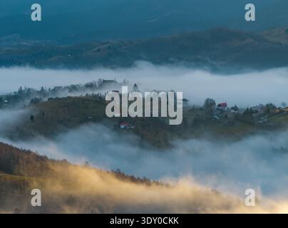 Vue aérienne du village niché parmi les collines, partiellement enveloppé dans la brume éthérée, baigné dans la douce lueur de l'aube, Șirnea, Brașov, Roumanie. Banque D'Images