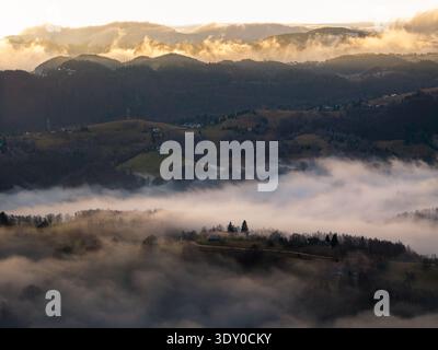 Vue aérienne de brume éthérée couvre les collines, berçant des villages lointains dans une douce étreinte sous un ciel doré, Șirnea, Brașov, Roumanie. Banque D'Images