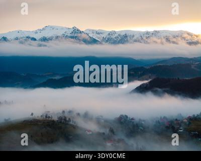 Vue aérienne d'un village niché dans une vallée recouverte d'un brouillard éthéré, avec des montagnes enneigées s'élevant majestueusement au loin, Șirnea, Brașov Banque D'Images