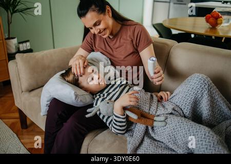 Mère attentionnée mesurant la température de son fils malade allongé sur le canapé Banque D'Images