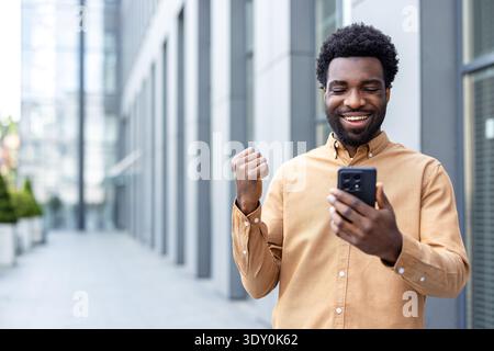 Homme souriant se sentant excité et faisant un geste de pompe du poing, célébrant les bonnes nouvelles ou les succès reçus sur son téléphone portable tout en se tenant à l'extérieur dans un cadre urbain Banque D'Images