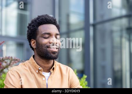 Jeune homme debout dehors avec les yeux fermés et un sourire serein, expérimentant un moment de calme et de pleine conscience, trouvant détente et paix intérieure en plein air Banque D'Images