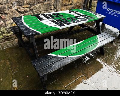 Édimbourg, Écosse, Royaume-Uni - 15 février 2026 : banc de pique-nique en bois imbibé de pluie avec logo vert audacieux contre un mur de pierre dans la cour d'un pub d'Édimbourg. Banque D'Images