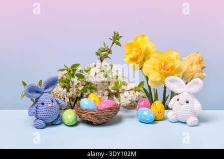 Lapins tricotés de Pâques blancs et bleus avec des œufs colorés dans un nid d'oiseau et des branches de cerises printanières en fleurs, des tulipes jaunes et des jonquilles sur fond bleu Banque D'Images