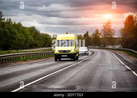 L'autobus scolaire jaune emprunte une route asphaltée humide. Voiture de tourisme blanche suit. Ciel couvert au-dessus de la tête. Arbres verts, ligne de feuillage route sinueuse. Garde-corps Alo Banque D'Images