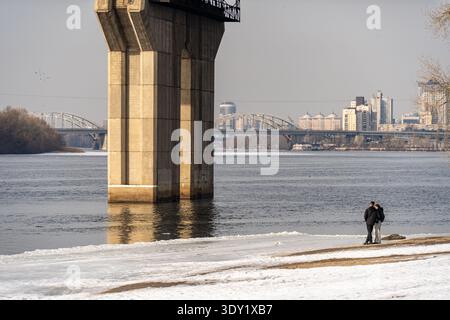 Vue d'un couple se dresse sur la rive enneigée sous un support de pont en béton, surplombant la rivière calme et la ligne d'horizon de la ville lointaine, Kiev, Kiev, Ukraine. Banque D'Images