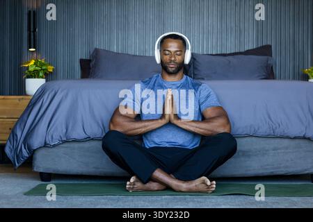 Homme afro-américain méditant sur un tapis de yoga dans la chambre portant une chemise athlétique bleue et des écouteurs. Pleine conscience, bien-être, calme, sérénité, minimaliste, Banque D'Images