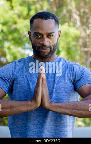 Homme afro-américain mature tenant les mains dans la prière à la poitrine à l'extérieur portant une chemise athlétique bleue. Pleine conscience, sérénité, portrait, parc, feuillage, s/o Banque D'Images