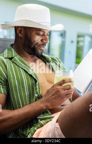 Homme afro-américain s'inclinant sur une chaise de patio dans une chemise rayée chapeau tenant un livret de lecture de limonade. Extérieur, terrasse, serein, ensoleillé, boisson, citru Banque D'Images