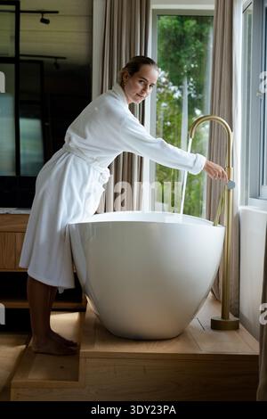 Femme dans la vingtaine en robe blanche appuyée sur les marches, actionnant le robinet d'or dans la baignoire remplissant la salle de bains. Baignoire, autoportante, bois, naturel, lumière, se Banque D'Images