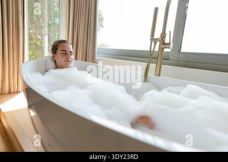 Femme couchée dans une baignoire blanche autoportante dans la salle de bains, entourée de mousse et robinet en laiton. Sérénité, spa, minimaliste, lumière du jour, fenêtre, oreillers, Banque D'Images