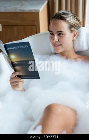 Femme couchée dans une baignoire blanche à la maison avec de la mousse, la tête sur la serviette tenant des couchers de soleil sans fin de livre de poche. Baignoire, bulles, lecture, autosoin, serein, ambiance Banque D'Images