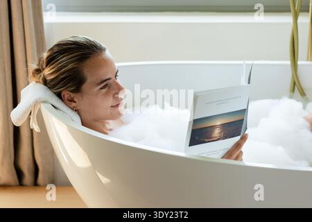 Femme s'inclinant dans une baignoire mousseuse autoportante, reposant la tête sur une serviette roulée et lisant le livre de poche. Salle de bain, spa, détente, roman, coucher de soleil, océan, pl Banque D'Images
