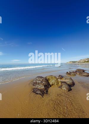 Plage de sable avec quelques gros rochers par une journée ensoleillée avec des vagues se lavant jusqu'au rivage Banque D'Images