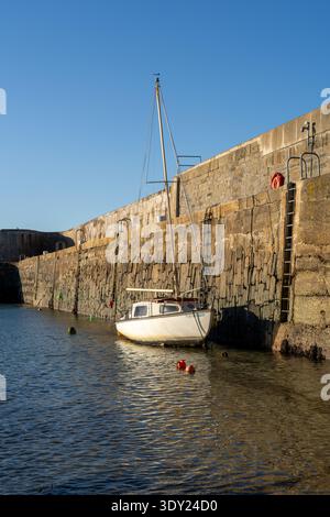 Un petit voilier blanc repose doucement amarré par des bouées contre le mur de pierre texturé du port à Portsoy. Banque D'Images