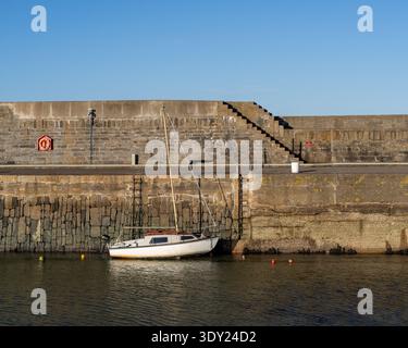 Un petit voilier blanc repose doucement amarré par des bouées contre le mur de pierre texturé du port à Portsoy. Banque D'Images