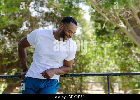 Homme africain mûr appuyé sur une balustrade en métal au parc, souriant, portant un t-shirt blanc et un short bleu. Nature, extérieur, bois, ensoleillé, feuillage, boke Banque D'Images