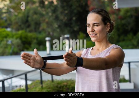 Femme dans les années 20 s'exerçant sur le balcon en utilisant la bande de résistance noire et portant un réservoir rose, espace de copie. Extérieur, fitness, entraînement, entraînement, bien-être, calme, Banque D'Images