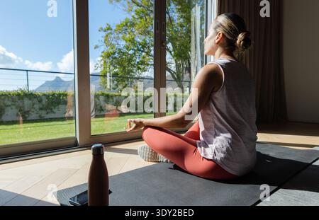 Femme adulte en leggings assis sur tapis par des portes coulissantes avec bouteille d'eau, téléphone, espace de copie. Yoga, bien-être, serein, ensoleillé, patio, jardin, nature Banque D'Images
