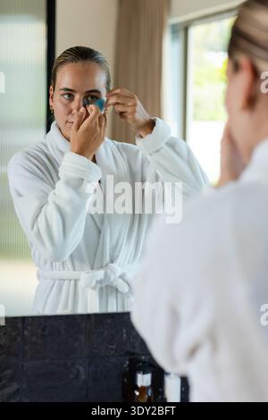 Femme portant un peignoir blanc à la vanité de la salle de bains, appliquant un patch de gel bleu sous les yeux dans le miroir. Femme, soins de la peau, autosoin, réflexion, lumière du jour, vent Banque D'Images