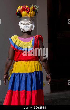 Femme afro-colombienne Palenquera portant une robe traditionnelle vibrante dans les couleurs jaune, bleu et rouge portant panier de fruits tropicaux équilibré sur elle HE Banque D'Images