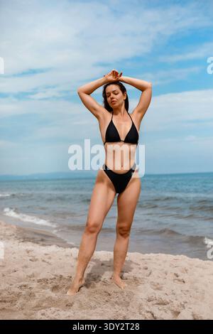 Jeune femme attrayante posant sur une plage de sable avec les mains au-dessus de la tête sous la lumière du soleil. Concept de protection solaire, soin UV de la peau et mode de vie sain Banque D'Images