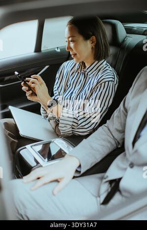 Une coréenne est assise dans une voiture concentrée sur son téléphone tandis qu'un homme caucasien est assis à côté d'elle. Ils sont tous les deux habillés dans des vêtements professionnels décontractés. Le CA Banque D'Images