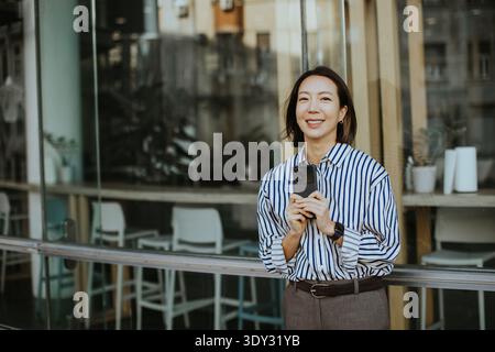 Une femme coréenne se tient devant un café tenant un smartphone. Elle sourit à la caméra en s'appuyant contre le mur de verre. Le cadre montre la vie urbaine Banque D'Images