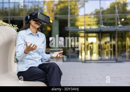 Femme d'affaires innovante portant un casque vr, faisant des gestes avec ses mains, profitant d'une simulation 3D ou d'une expérience de jeu immersive tout en étant assise sur un banc extérieur dans un environnement urbain Banque D'Images