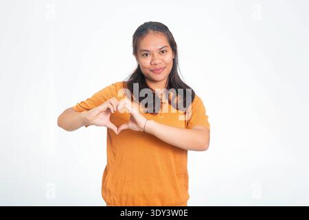 Heureuse femme indonésienne en chemise orange créant un symbole de coeur avec ses doigts, souriant chaleureusement sur un fond blanc. Banque D'Images