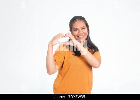 Heureuse femme indonésienne en chemise orange créant un symbole de coeur avec ses doigts, souriant chaleureusement sur un fond blanc. Banque D'Images