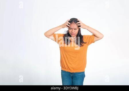 Jeune femme indonésienne en chemise orange paraissant stressée et souffrant d'une migraine ou de maux de tête, touchant sa tête dans la douleur. Banque D'Images