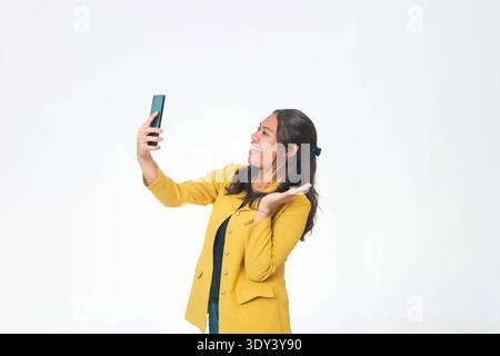 Vue latérale de la femme indonésienne souriante en blazer jaune tenant smartphone pour selfie avec geste de paume ouvert, regardant le téléphone, fond blanc. Banque D'Images