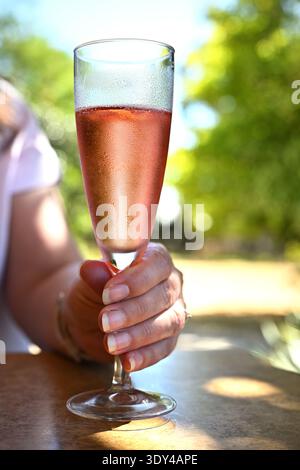 Gros plan de la main tenant un verre de vin mousseux rosé frais avec condensation sur le verre au café extérieur avec fond bokeh vert doux d'été Banque D'Images