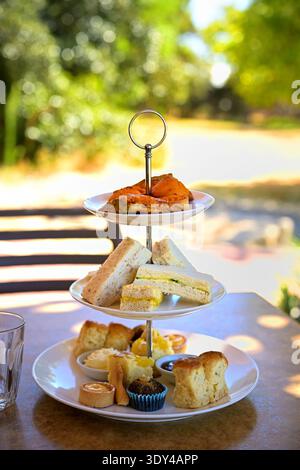 Stand de thé de l'après-midi à trois niveaux avec sandwichs, scones, pâtisseries et gâteaux sur la table de café extérieure avec fond de jardin bokeh vert doux Banque D'Images