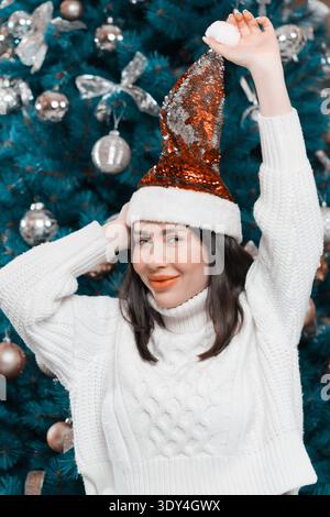 Une jeune femme heureuse aux yeux bleus sourit tout en portant un chapeau de Père Noël à sequins festif et un pull blanc confortable. Mannequin pose ludique devant un décor Banque D'Images