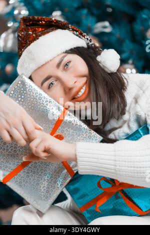 Portrait d'une jeune femme heureuse dans un chapeau de Père Noël embrassant des cadeaux de Noël. Dame sourit, tenant des cadeaux en argent et bleu emballage avec des rubans rouges. Fe Banque D'Images