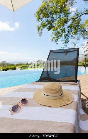 Chapeau de soleil en paille tissé assis sur une serviette rayée sur une terrasse en bois, avec lunettes de soleil et piscine à débordement Banque D'Images