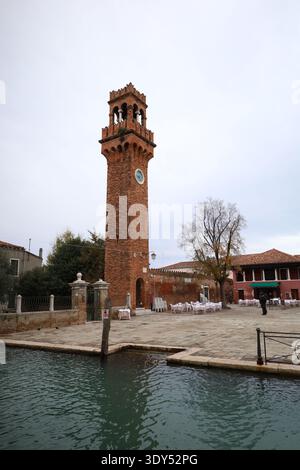 Un grand clocher en briques s'élève à côté d'un canal, avec une place ouverte avec des tables et des chaises disposées, bâtiments à proximité. Banque D'Images