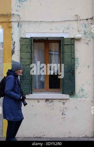 Individu portant un manteau et un chapeau marche à côté d'un mur de plâtre usé avec de la peinture pelable et des volets en bois vert encadrant une petite fenêtre. Banque D'Images
