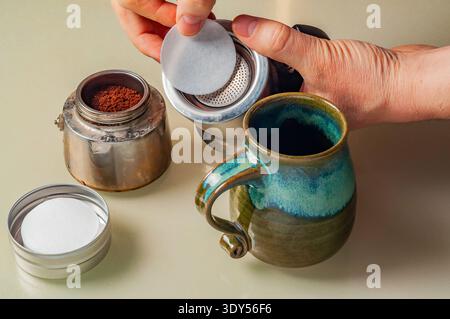 L'image montre une personne préparant le café avec un pot Moka, plaçant un papier filtre. Une tasse en céramique complète la préparation confortable du café. Banque D'Images